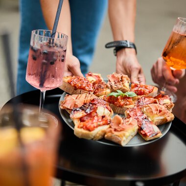 A tray with antipasti, garnished with ham and tomatoes. Surrounded by drinks and people enjoying a sociable time. | © Kleinwalsertal Tourismus | Oliver Farys