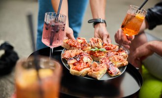 A tray with antipasti, garnished with ham and tomatoes. Surrounded by drinks and people enjoying a sociable time. | © Kleinwalsertal Tourismus | Oliver Farys