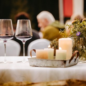 A beautifully set table with two wine glasses and rustic decor. In the background, guests can be seen at an event. | © Kleinwalsertal Tourismus eGen | Oliver Farys