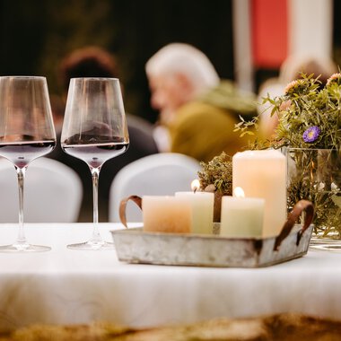 Two wine glasses are placed on a table, surrounded by candles and a small floral decoration. In the background, blurry guests can be seen at a celebration. | © Kleinwalsertal Tourismus eGen | Oliver Farys
