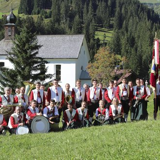 A traditional costumes band stands on a meadow in front of a small church. In the background, green hills and trees can be seen. | © Trachtenkapelle Harmonie Mittelberg | Jockl Metzger