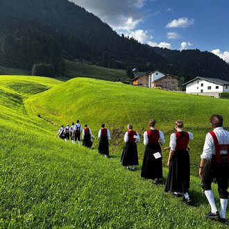 A traditional brass band in traditional costumes stands on a meadow in front of a rustic wooden house. The musicians hold their instruments and smile at the camera, with mountains in the background. | © Trachtenkapelle Riezlern