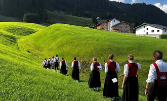 A traditional brass band in traditional costumes stands on a meadow in front of a rustic wooden house. The musicians hold their instruments and smile at the camera, with mountains in the background. | © Trachtenkapelle Riezlern