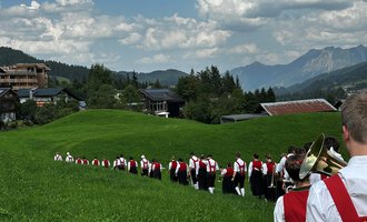 A group of people in traditional costumes is hiking across a green meadow. In the background, mountains and a clear sky can be seen. | © Trachtenkapelle Riezlern