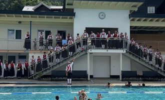 A swimming pool with many people standing on the terrace and watching. In the background, mountains and a blue sky can be seen. | © Trachtenkapelle Riezlern
