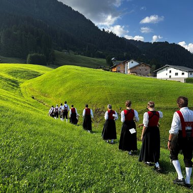 A traditional brass band in traditional costumes stands on a meadow in front of a rustic wooden house. The musicians hold their instruments and smile at the camera, with mountains in the background. | © Trachtenkapelle Riezlern