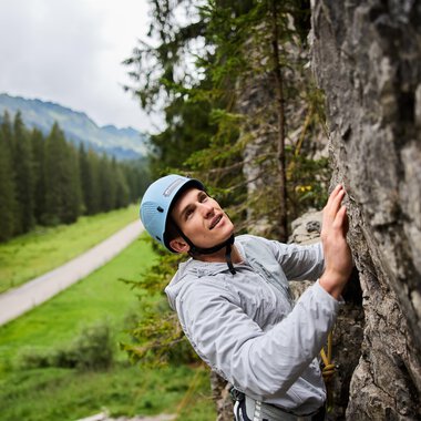 Ein Kletterer, der an einer Felswand arbeitet, trägt einen Helm und konzentriert sich auf seinen Aufstieg. Im Hintergrund sind Bäume und eine Straße in einer grünen Landschaft zu sehen. | © Bergschule Kleinwalsertal | Oliver Farys