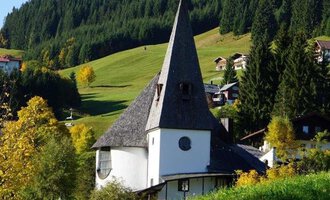 A picturesque church with a tall, pointed tower amidst a green landscape. In the background, gentle hills and some traditional houses are visible. | © Ev. Kirche Kleinwalsertal