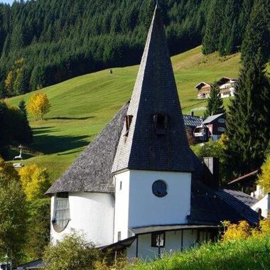 A picturesque church with a tall, pointed tower amidst a green landscape. In the background, gentle hills and some traditional houses are visible. | © Ev. Kirche Kleinwalsertal