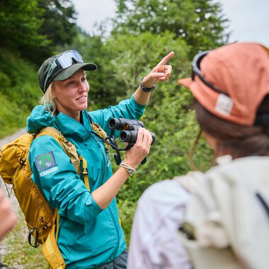 Eine Frau mit einer Kamera erklärt etwas einer Gruppe von Personen im Freien. Im Hintergrund sind Bäume und eine hügelige Landschaft zu sehen. | © Kleinwalsertal Tourismus eGen | Oliver Farys