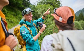 Eine Frau mit einer Kamera erklärt etwas einer Gruppe von Personen im Freien. Im Hintergrund sind Bäume und eine hügelige Landschaft zu sehen. | © Kleinwalsertal Tourismus eGen | Oliver Farys