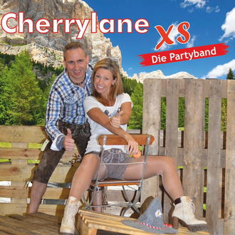 Ein Paar posiert auf einer rustikalen Holzbank vor einer schönen Berglandschaft. Sie tragen traditionelle Kleidung und lächeln fröhlich in die Kamera.