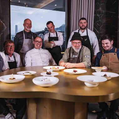 A group of chefs is sitting at a round table with several elegant plates. In the background, there are large windows with a view of the mountains. | © Frank Simon