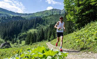 A runner jogs on a narrow path in the mountains. The landscape is green with beautiful forests and tall mountains in the background. | © Kleinwalsertal Tourismus - Dominik Berchtold