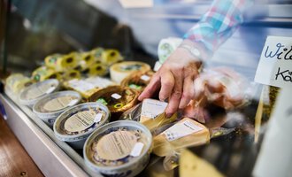 Ein Verkaufsstand mit verschiedenen Käsesorten. Eine Hand greift nach einem Käseprodukt in der Auslage. | © Kleinwalsertal Tourismus | Fotograf Oliver Farys