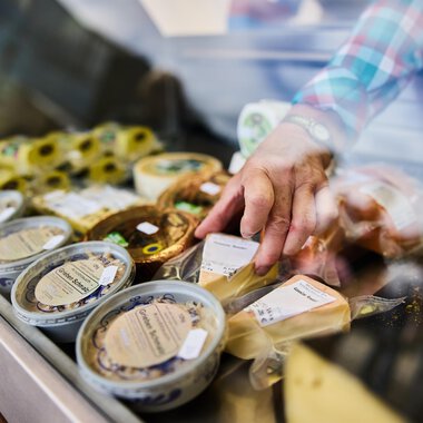 A sales stand with various types of cheese. A hand reaches for a cheese product in the display. | © Kleinwalsertal Tourismus | Fotograf Oliver Farys