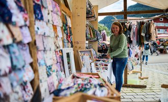 A market stand with various colorful fabrics and clothing. A woman is browsing the displays and enjoying the atmosphere. | © Kleinwalsertal Tourismus | Fotograf Oliver Farys