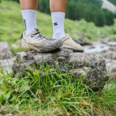 A pair of feet is standing on a large stone in a green, natural area. In the background, trees and a small river are visible.