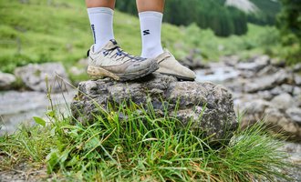 A pair of feet is standing on a large stone in a green, natural area. In the background, trees and a small river are visible.