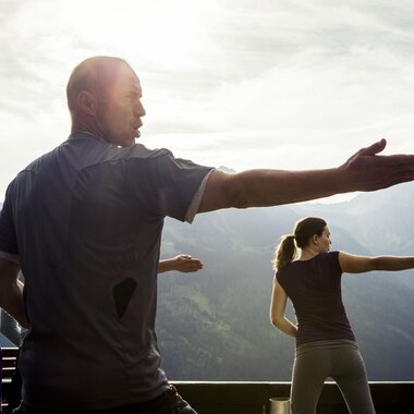 A group of people is practicing Tai Chi in nature. In the background, majestic mountains and a slightly cloudy sky are visible. | © Kleinwalsertal Tourismus | Oliver Farys