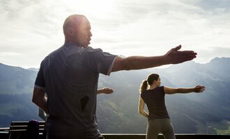 A group of people is practicing Tai Chi in nature. In the background, majestic mountains and a slightly cloudy sky are visible. | © Kleinwalsertal Tourismus | Oliver Farys