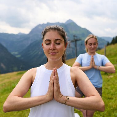 Two women are practicing yoga in the mountains. The surroundings are green and impressive, featuring tall mountains and a cloudy sky. | © Kleinwalsertal Tourismus | Oliver Farys
