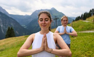 Two women are practicing yoga in the mountains. The surroundings are green and impressive, featuring tall mountains and a cloudy sky. | © Kleinwalsertal Tourismus | Oliver Farys
