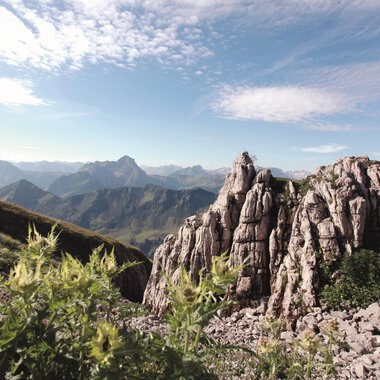 An impressive mountain landscape with rugged rocks and majestic mountains in the background. The sky is clear and the surroundings are characterized by green vegetation. | © Kleinwalsertal Tourismus eGen | Frank Drechsel