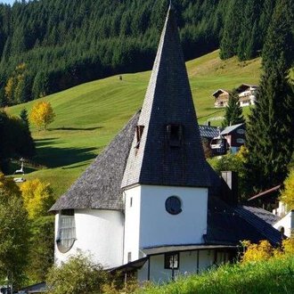 A traditional building with a tall pointed roof stands in a picturesque landscape.  
Surrounded by green hills and colorful trees, the scene appears peaceful and inviting. | © Evangelische Kreuzkirche Hirschegg