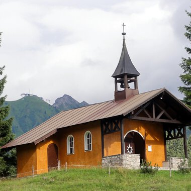 A small, charming church in the mountains. It has a pointed tower and is surrounded by green trees. | © Bruder Klaus Kapelle | Monika Gmeiner