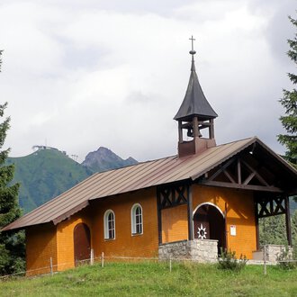 A small, charming church in the mountains. It has a pointed tower and is surrounded by green trees. | © Bruder Klaus Kapelle | Monika Gmeiner
