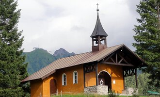 A small, charming church in the mountains. It has a pointed tower and is surrounded by green trees. | © Bruder Klaus Kapelle | Monika Gmeiner