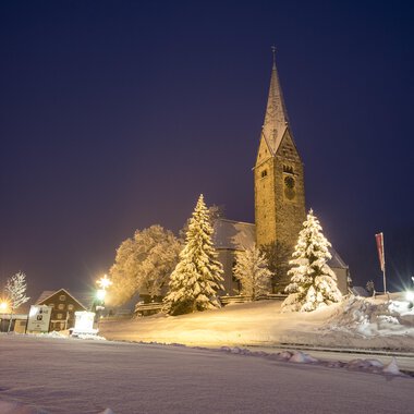 Eine verschneite Landschaft bei Nacht mit einer Kirche und beleuchteten Bäumen. Der Himmel ist dunkelblau und die Straße ist sichtbar. | © Kleinwalsertal Tourismus | Dominik Berchtold
