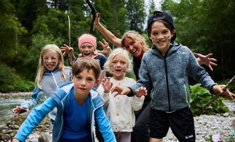 A group of children and an adult standing at the riverbank. They are having fun and posing with happy facial expressions. | © Kleinwalsertal Tourismus | Oliver Farys