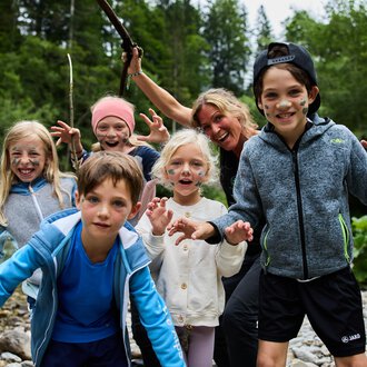 Eine Gruppe von Kindern und einem Erwachsenen, die am Ufer eines Flusses stehen. Sie haben Spaß und posieren mit fröhlichen Gesichtsausdrücken. | © Kleinwalsertal Tourismus | Oliver Farys