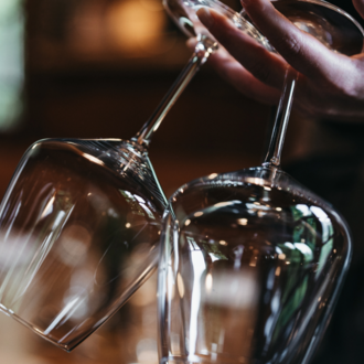 A person is holding two elegant wine glasses. The background is blurred, creating a cozy atmosphere. | © Winzer zu Tisch | Naturhotel Chesa Valisa