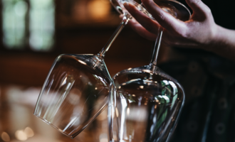 A person is holding two elegant wine glasses. The background is blurred, creating a cozy atmosphere. | © Winzer zu Tisch | Naturhotel Chesa Valisa