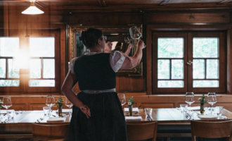A waitress in traditional attire stands with a glass in hand in a cozy restaurant. The wooden space is furnished with tables and stylish windows. | © Winzer zu Tisch | Naturhotel Chesa Valisa