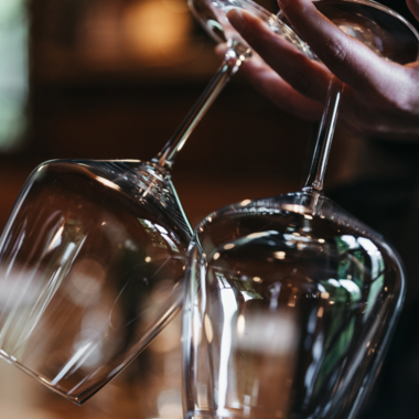 A person is holding two elegant wine glasses. The background is blurred, creating a cozy atmosphere. | © Winzer zu Tisch | Naturhotel Chesa Valisa