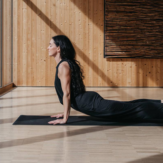 A bright yoga room with wooden paneling and large windows. Three people are practicing yoga and looking outside. | © Naturhotel Chesa Valisa