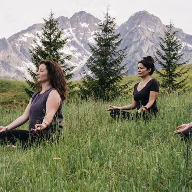 Ein heller Yogaraum mit Holzverkleidung und großen Fenstern. Drei Personen praktizieren Yoga und blicken nach draußen. | © Naturhotel Chesa Valisa
