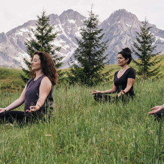 A bright yoga room with wooden paneling and large windows. Three people are practicing yoga and looking outside. | © Naturhotel Chesa Valisa