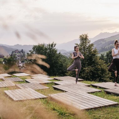 Zwei Frauen üben Yoga auf Holzplattformen in einer grünen Wiese. Im Hintergrund sind Berge und eine malerische Landschaft zu sehen.