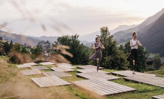 Zwei Frauen üben Yoga auf Holzplattformen in einer grünen Wiese. Im Hintergrund sind Berge und eine malerische Landschaft zu sehen.
