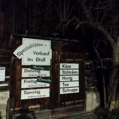 Ein Holzgebäude mit einem Verkaufsschild für Spezialitäten im Stall. Die Beschilderung weist auf Öffnungszeiten und angebotene Produkte wie Käse und Honig hin. | © Abler Spezialitätenverkauf im Stall | N. Lughammer