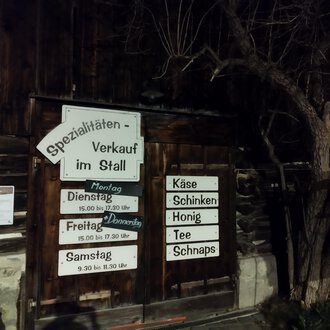 Ein Holzgebäude mit einem Verkaufsschild für Spezialitäten im Stall. Die Beschilderung weist auf Öffnungszeiten und angebotene Produkte wie Käse und Honig hin. | © Abler Spezialitätenverkauf im Stall | N. Lughammer