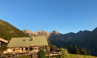 Eine malerische Almhütte umgeben von Bergen. Der klare Himmel und die grüne Wiese verleihen der Szene eine friedliche Atmosphäre. | © Alpe Kuhgehren | Lisa Hiesinger