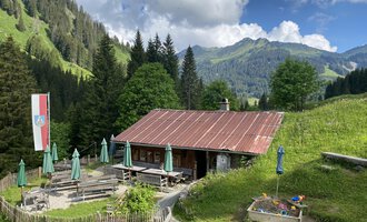 Eine gemütliche Berghütte mit rotem Dach und grünen Sonnenschirmen. Im Hintergrund sind grüne Hügel und Berge unter einem klaren Himmel zu sehen. | © Alpe Widderstein | Tom Egger