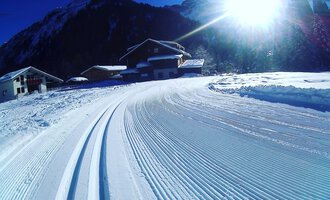 Eine verschneite Landschaft mit gut präparierten Loipe. Im Hintergrund sind Berge und chalets zu sehen, während die Sonne hell scheint.