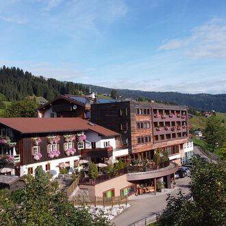 A large hotel building in a rural setting with green hills in the background. The sky is clear and the landscape looks inviting. | © Das Alphotel Hirschegg Hotel GmbH | Mark Kerwell